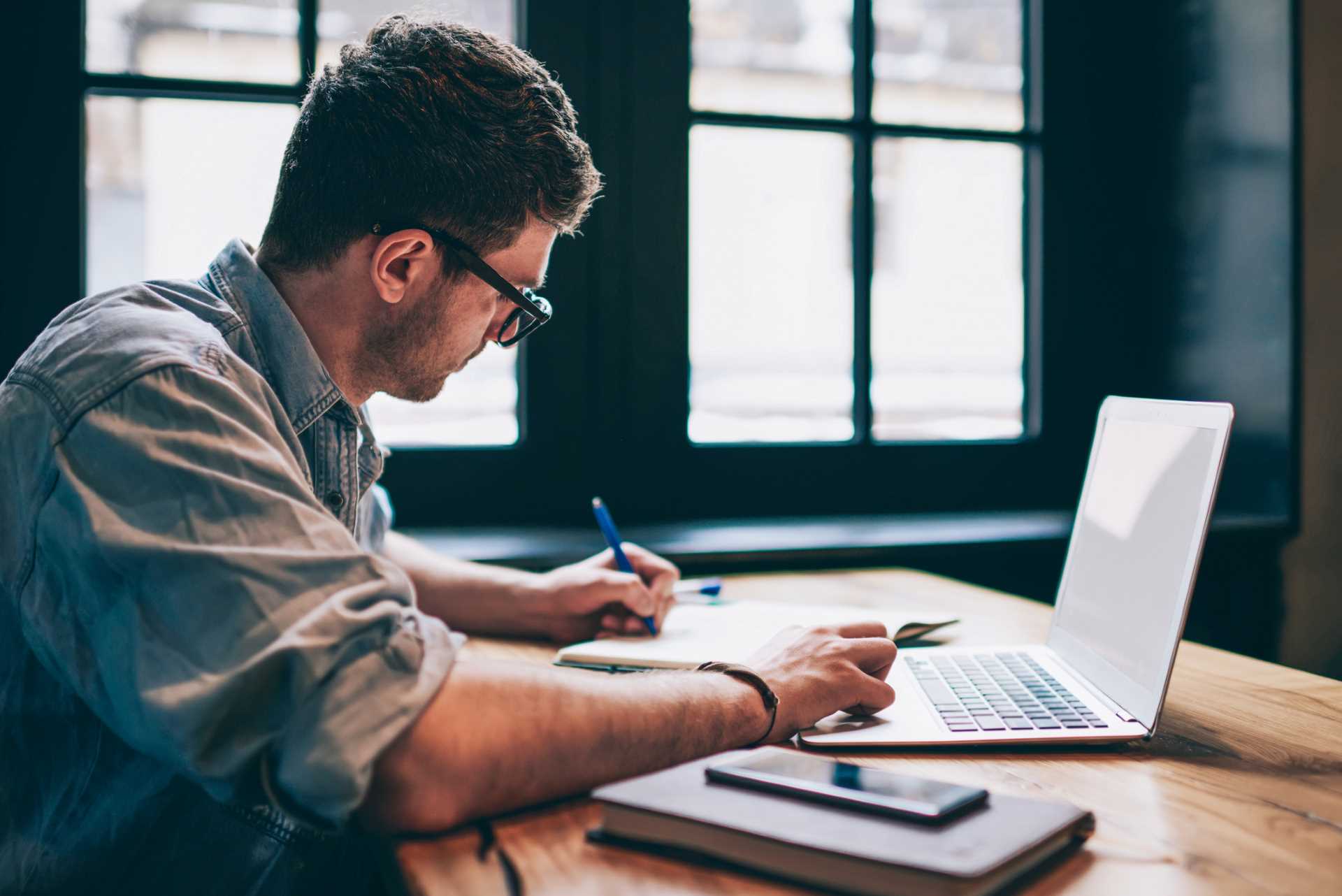 young man writes in a notebook sitting at a desk with laptop