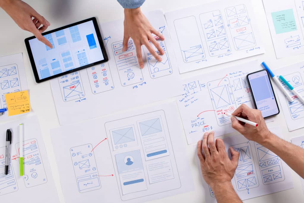 UX/UI designers discussing and brainstorming on wireframes for a website and mobile app prototype, surrounded by sketches of user flow and design tools.